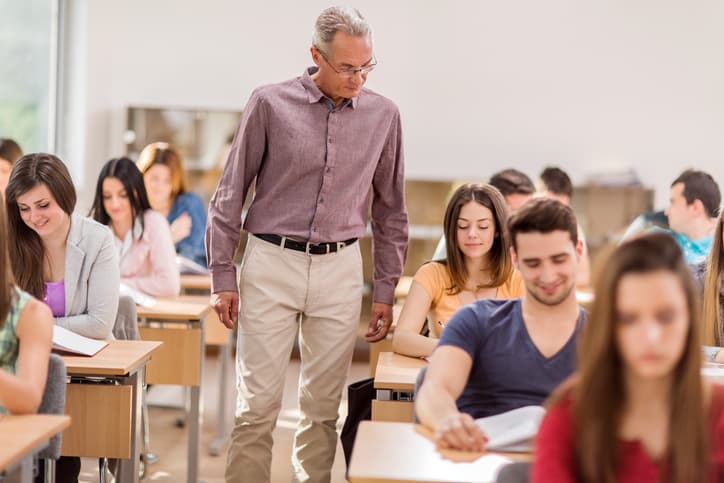 Large group of college students doing an exam in the classroom. Mature teacher is walking among them and monitoring.