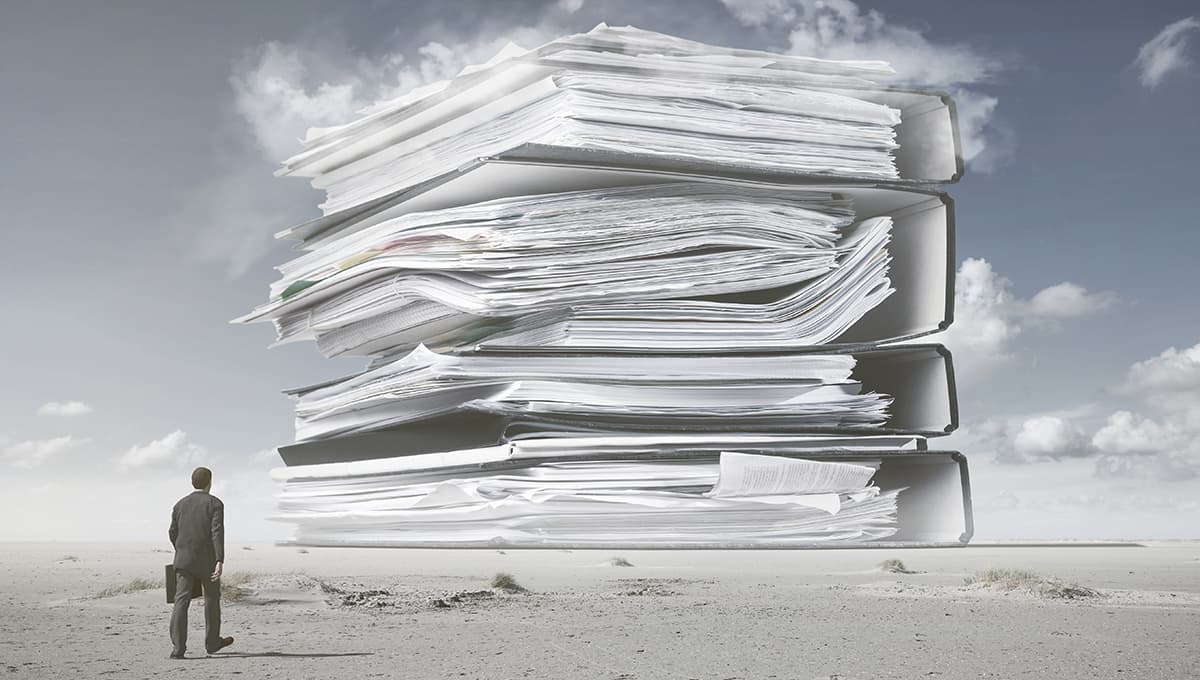 A man in a suit carrying a briefcase looks up in the desert at towering ring binders of paperwork