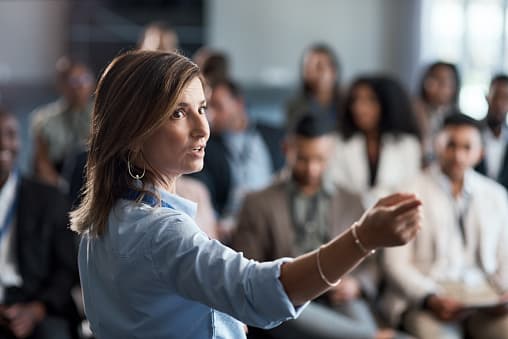 A woman at a business conference speaks and points to something off screen