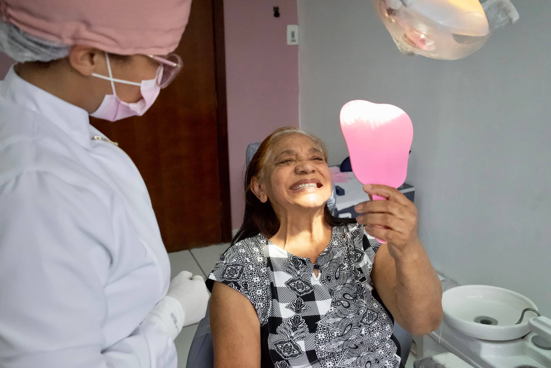 Image of a person sitting in a dental office holding a mirror up to see their teeth.