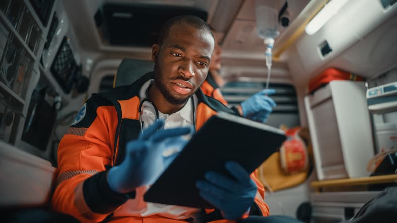 Black EMS Professional Paramedic Using Tablet Computer to Fill a Questionnaire for the Injured Patient on the Way to Hospital. Emergency Care Assistant Comforting the Patient in an Ambulance.