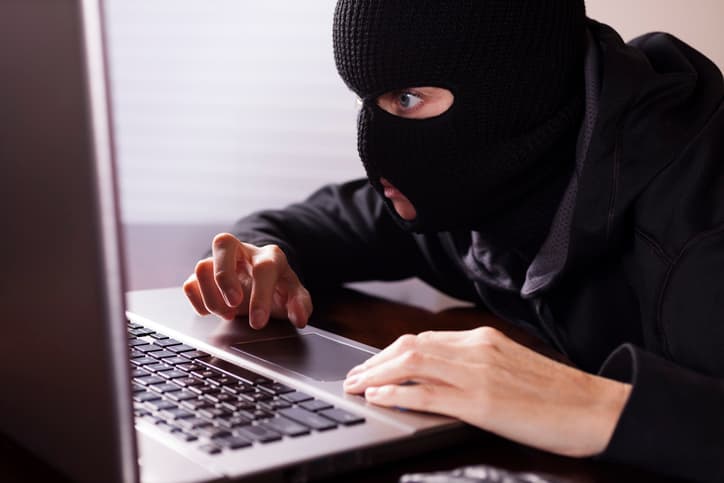 Masked theif wearing all black steals computer secrets or commits identity theft on an office or home laptop computer. Cyper crimes concept.