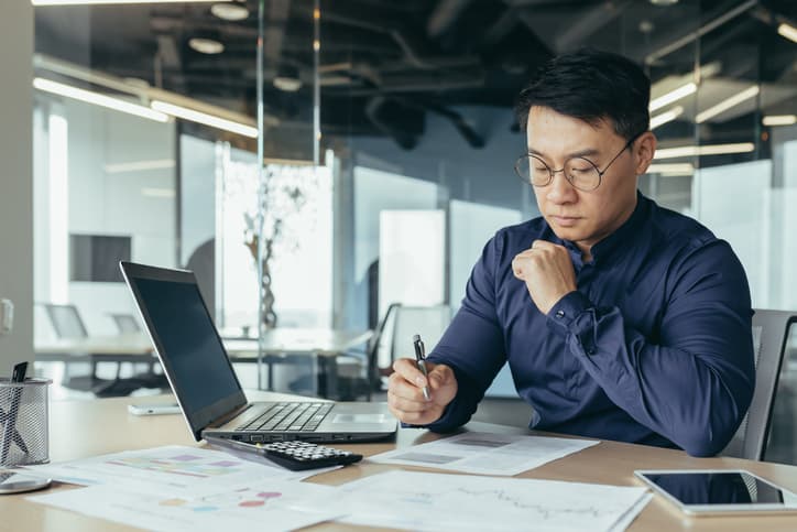 Thinking asian financier in glasses accountant working with documents and accounts, using laptop, male businessman doing paperwork in sitting at desk inside office building.