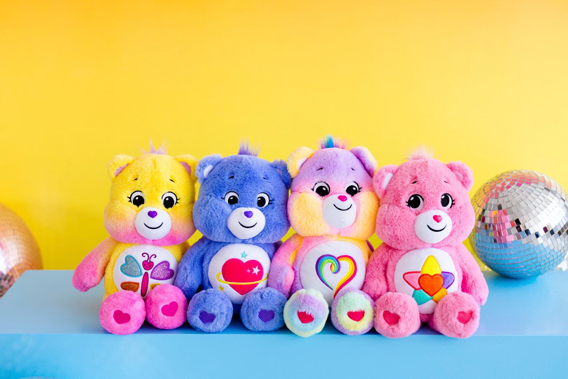 pink, blue and lavendar Care Bear stuffed animals sitting in front of yellow background