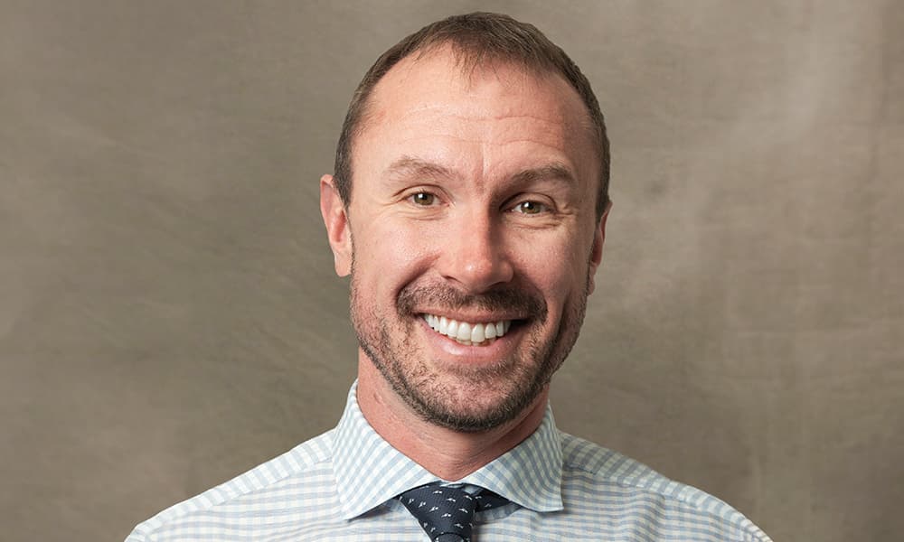 Headshot of John Stecher in a button-down shirt and tie against a beige background