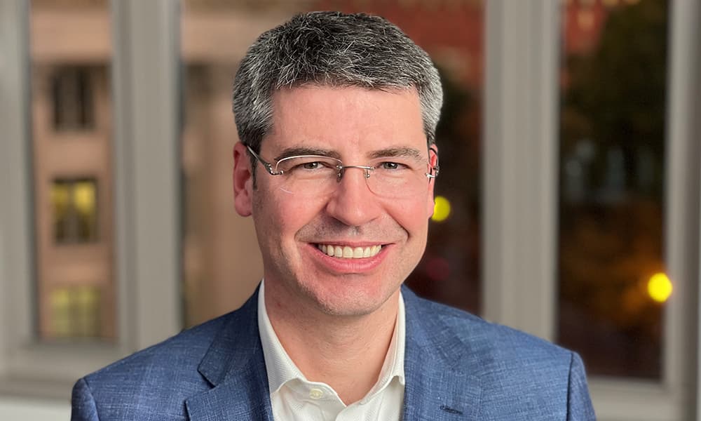 Headshot of Jay Scanlan in blue blazer and office buildings behind