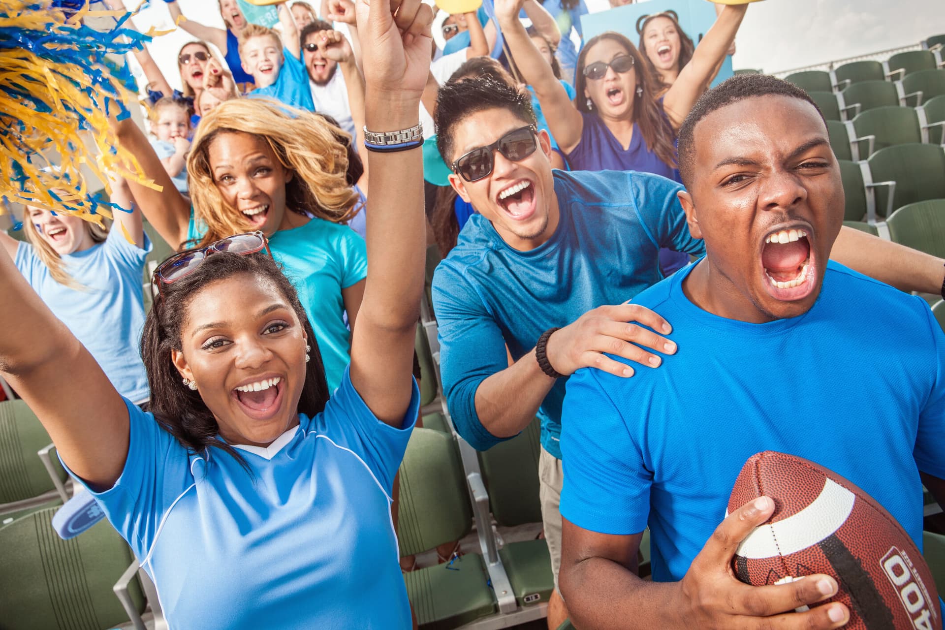 A large group of young adult football fans in outdoor stadium seats and dressed in blue clothing yell and cheer with excitement for their team.