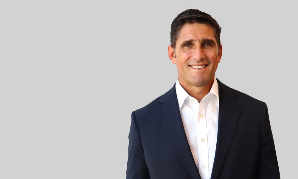 Fraser van Rensburg headshot in a white shirt and black blazer against a gray bacground.