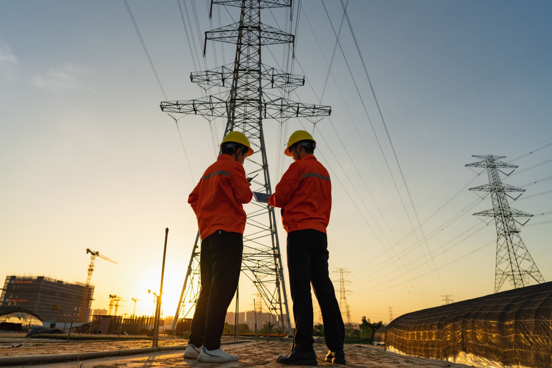 Two electric workers using a tablet to record and check the transmission line at sunset.