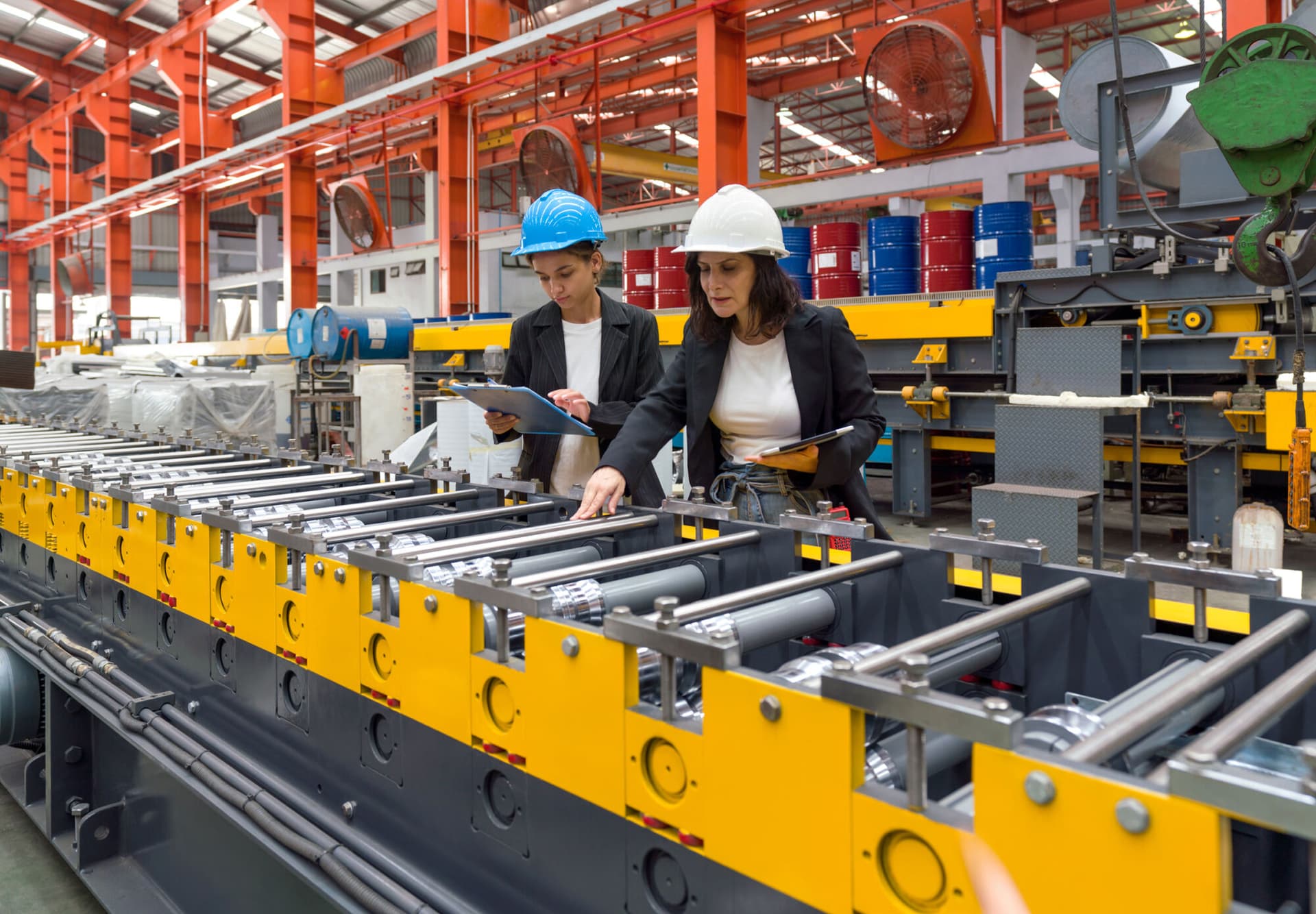 Two women operate machinery in a factory, wearing safety gear and focused on their task.