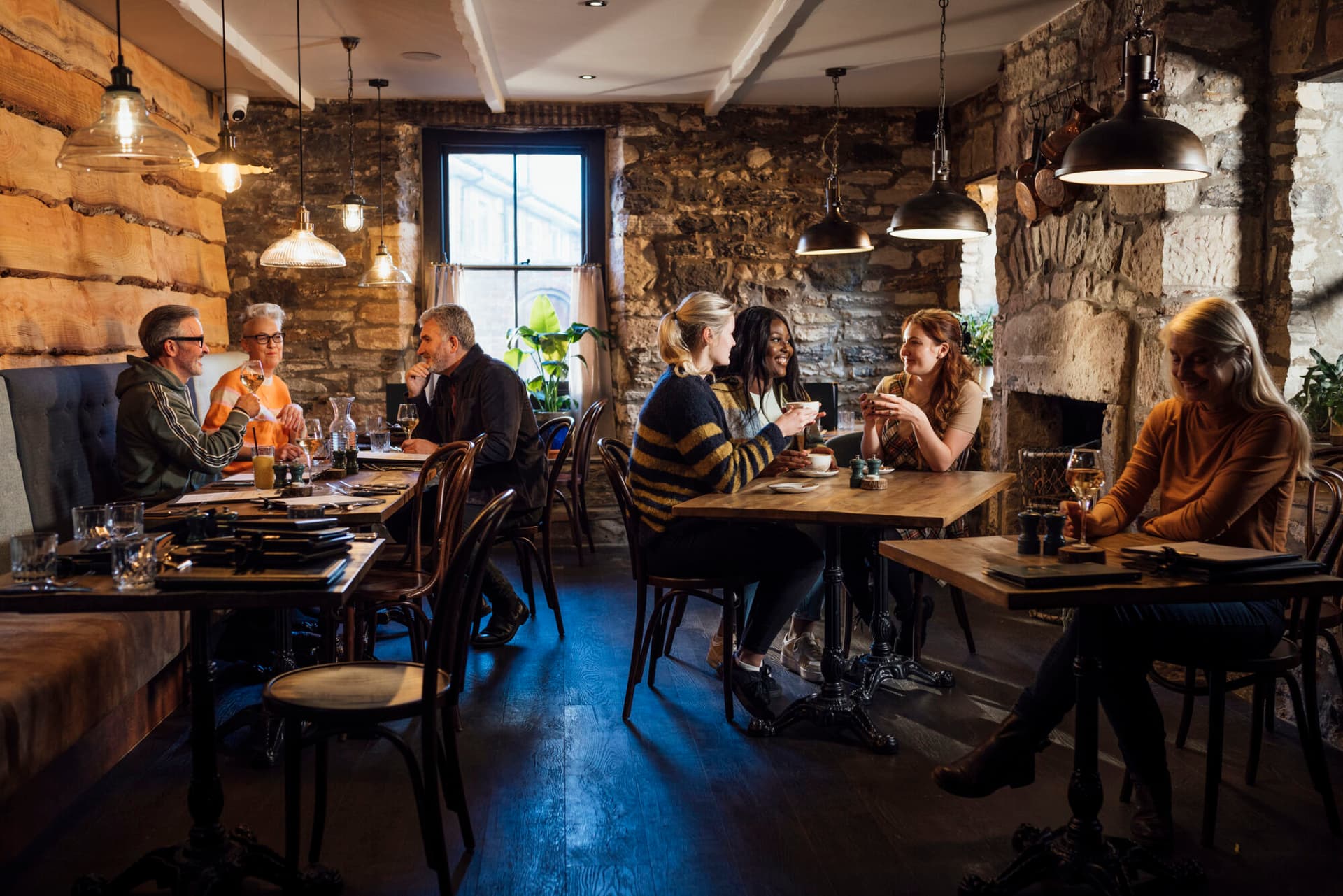 A wide angle view of a group of diners in their friendship groups enjoying a meal in a warm coastal pub in Seahouses in the North East of England. They are sat inside the warm glass conservatory area of the pub in the dining area and enjoying some rustic food and drink
