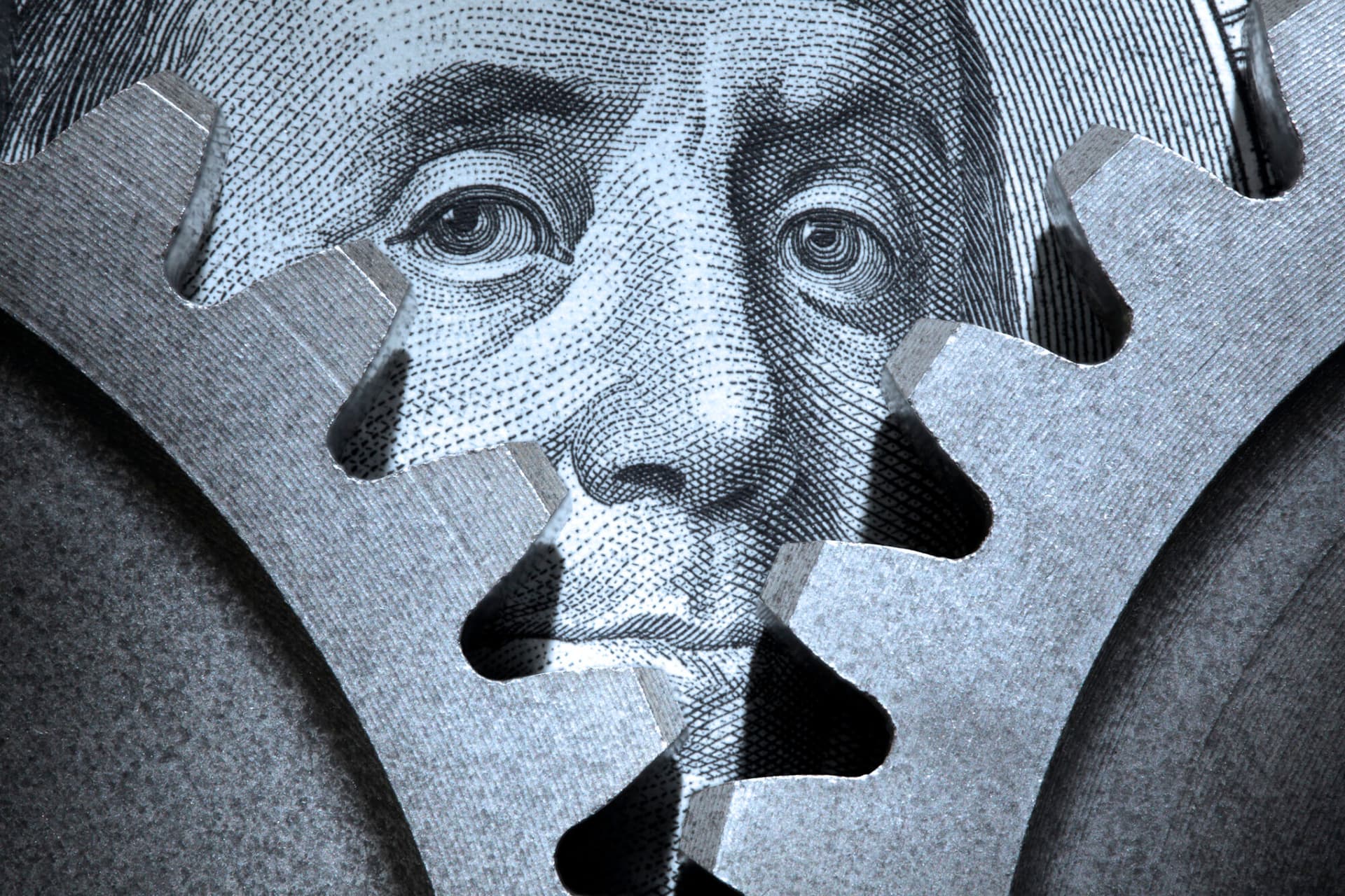 A pair of interlocking gears on a $100 bill with the portrait of Benjamin Franklin peering out