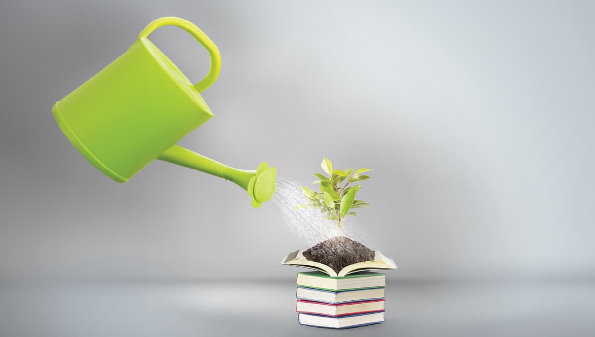 A watering can pouring water over a pile of books with one open at the top and a plant growing out of it